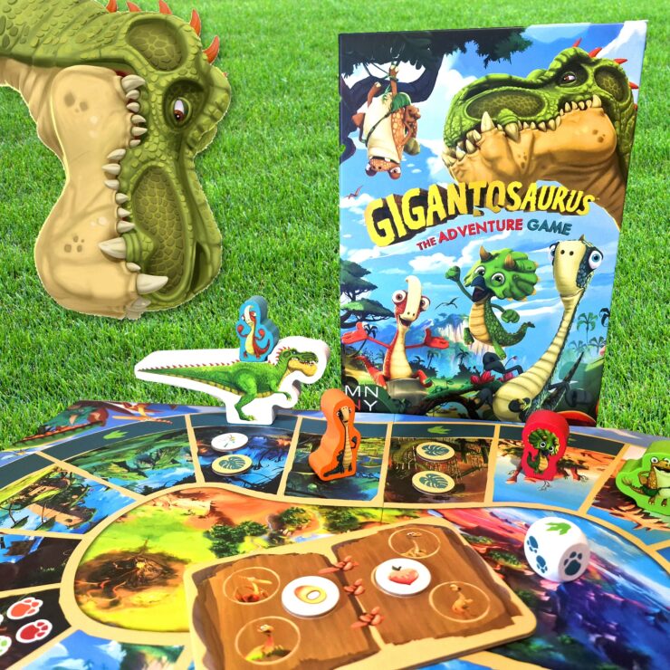 Colorful Gigantosaurus board game setup featuring a dinosaur-themed game board, character pieces, and the game box, all displayed on green grass. Perfect for family game nights and children's entertainment.