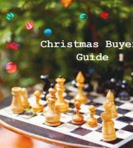 Chessboard with wooden pieces in front of a decorated Christmas tree, featuring a text overlay that reads "Christmas Buyers Guide." Ideal for holiday gift ideas related to chess and games.