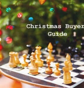 Chessboard with wooden pieces in front of a decorated Christmas tree, featuring a text overlay that reads "Christmas Buyers Guide." Ideal for holiday gift ideas related to chess and games.