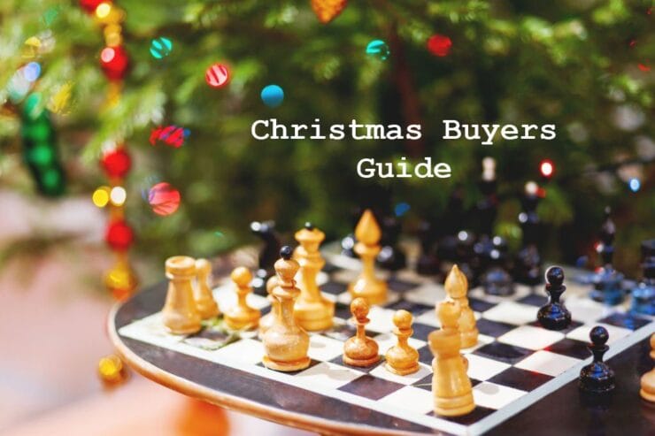 Chessboard with wooden pieces in front of a decorated Christmas tree, featuring a text overlay that reads "Christmas Buyers Guide." Ideal for holiday gift ideas related to chess and games.