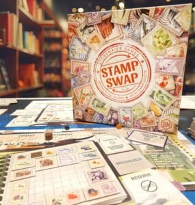 Board game "Stamp Swap" by Stonemaier Games displayed on a table, featuring colorful game components, including a game board, cards, and tokens, with a bookshelf in the background.