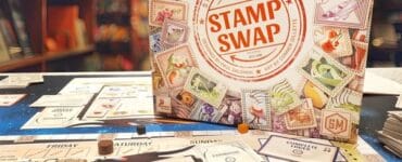 Board game "Stamp Swap" by Stonemaier Games displayed on a table, featuring colorful game components, including a game board, cards, and tokens, with a bookshelf in the background.