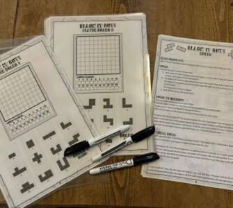 Printable game sheets for "Black It Out!" featuring player boards and game rules. The image showcases two player boards with grids and shapes, along with a rules sheet explaining gameplay and objectives. Markers are placed beside the sheets, ready for use.