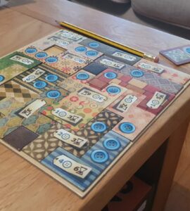 Colorful board game setup on a wooden table, featuring a patterned game board with blue buttons and numbered tiles. A yellow pencil is placed beside the board, with a small card showing a blue token in the corner. The background includes a soft couch, creating a cozy gaming atmosphere.