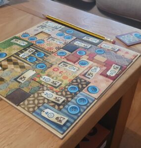 Colorful board game setup on a wooden table, featuring a patterned game board with blue buttons and numbered tiles. A yellow pencil is placed beside the board, with a small card showing a blue token in the corner. The background includes a soft couch, creating a cozy gaming atmosphere.