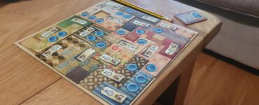 Colorful board game setup on a wooden table, featuring a patterned game board with blue buttons and numbered tiles. A yellow pencil is placed beside the board, with a small card showing a blue token in the corner. The background includes a soft couch, creating a cozy gaming atmosphere.
