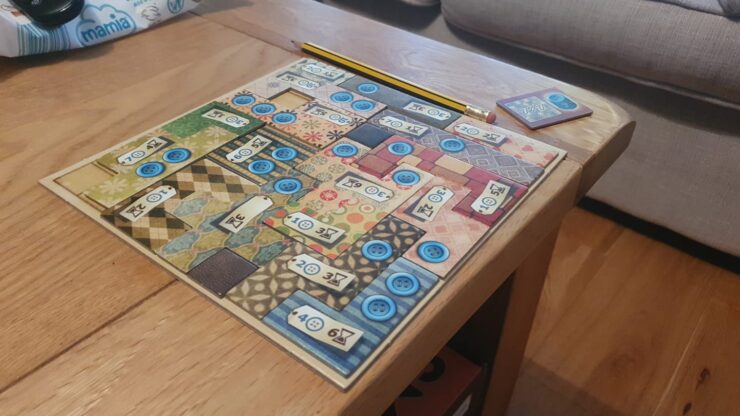 Colorful board game setup on a wooden table, featuring a patterned game board with blue buttons and numbered tiles. A yellow pencil is placed beside the board, with a small card showing a blue token in the corner. The background includes a soft couch, creating a cozy gaming atmosphere.