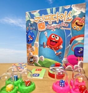 Colorful board game "Jumping Dice" featuring cartoon dice characters, game components including dice, cards, and a bell, set against a blue sky background.