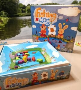 Colorful board game "Fishing Day" featuring a playful fishing scene, with vibrant wooden pieces and a scenic backdrop of a river and greenery. Ideal for family fun and skill development.