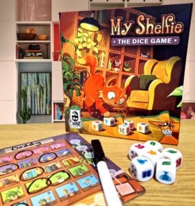 Image of the board game "My Shelfie: The Dice Game" featuring a playful cat on the box cover, alongside game components including dice and a score sheet. The background shows a modern living space with bookshelves and decor, creating a cozy atmosphere for family game night.