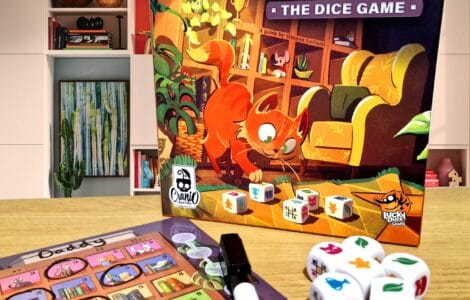 Image of the board game "My Shelfie: The Dice Game" featuring a playful cat on the box cover, alongside game components including dice and a score sheet. The background shows a modern living space with bookshelves and decor, creating a cozy atmosphere for family game night.