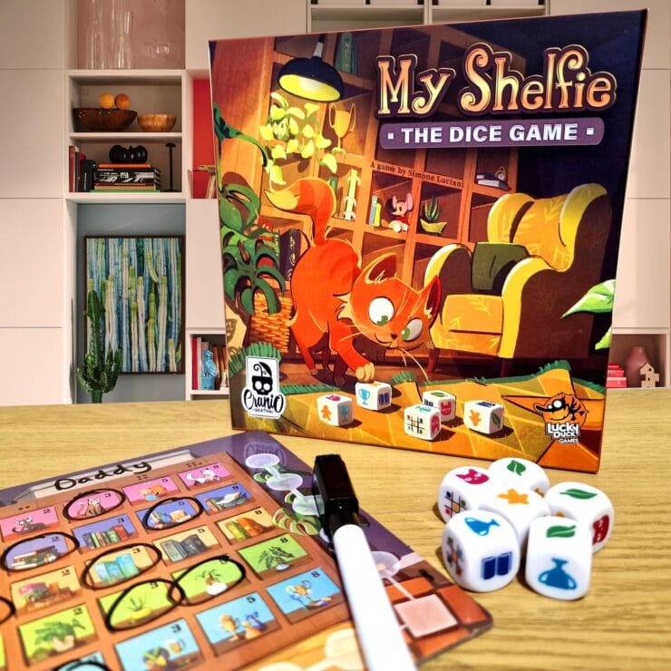 Image of the board game "My Shelfie: The Dice Game" featuring a playful cat on the box cover, alongside game components including dice and a score sheet. The background shows a modern living space with bookshelves and decor, creating a cozy atmosphere for family game night.