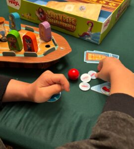 Child's hands engaged in gameplay with colorful game pieces on a green table, featuring the HABA board game "Captain Pepe: Treasure Ahoy!" The game components include a boat, hourglass, and various tokens.