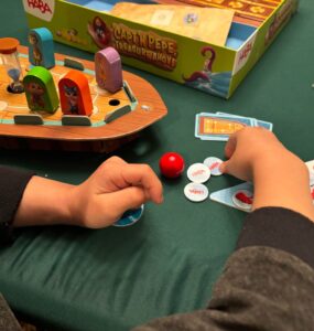 Child's hands engaged in gameplay with colorful game pieces on a green table, featuring the HABA board game "Captain Pepe: Treasure Ahoy!" The game components include a boat, hourglass, and various tokens.