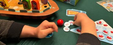 Child's hands engaged in gameplay with colorful game pieces on a green table, featuring the HABA board game "Captain Pepe: Treasure Ahoy!" The game components include a boat, hourglass, and various tokens.