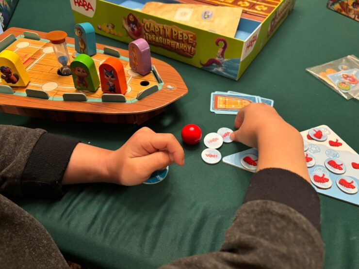 Child's hands engaged in gameplay with colorful game pieces on a green table, featuring the HABA board game "Captain Pepe: Treasure Ahoy!" The game components include a boat, hourglass, and various tokens.