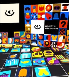 Colorful game pieces and the box of "Wilmot's Warehouse" are displayed against a black background. The box features a playful design with various shapes and a smiling face, while the scattered game pieces include vibrant icons in different colors and styles, showcasing the game's artistic theme.