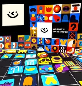 Colorful game pieces and the box of "Wilmot's Warehouse" are displayed against a black background. The box features a playful design with various shapes and a smiling face, while the scattered game pieces include vibrant icons in different colors and styles, showcasing the game's artistic theme.