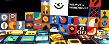 Colorful game pieces and the box of "Wilmot's Warehouse" are displayed against a black background. The box features a playful design with various shapes and a smiling face, while the scattered game pieces include vibrant icons in different colors and styles, showcasing the game's artistic theme.
