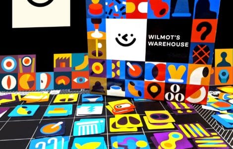 Colorful game pieces and the box of "Wilmot's Warehouse" are displayed against a black background. The box features a playful design with various shapes and a smiling face, while the scattered game pieces include vibrant icons in different colors and styles, showcasing the game's artistic theme.