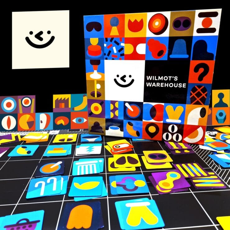 Colorful game pieces and the box of "Wilmot's Warehouse" are displayed against a black background. The box features a playful design with various shapes and a smiling face, while the scattered game pieces include vibrant icons in different colors and styles, showcasing the game's artistic theme.