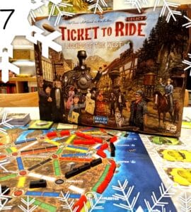 Alt text: Ticket to Ride: Legends of the West board game box displayed on a table with an illustrated game board featuring colorful train routes and player pieces. The scene is decorated with snowflake graphics, indicating a festive theme.