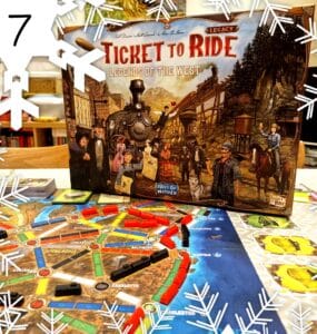 Alt text: Ticket to Ride: Legends of the West board game box displayed on a table with an illustrated game board featuring colorful train routes and player pieces. The scene is decorated with snowflake graphics, indicating a festive theme.