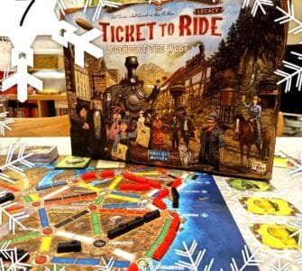 Alt text: Ticket to Ride: Legends of the West board game box displayed on a table with an illustrated game board featuring colorful train routes and player pieces. The scene is decorated with snowflake graphics, indicating a festive theme.