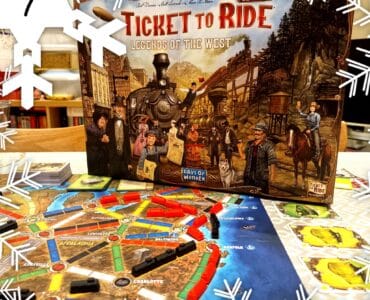 Alt text: Ticket to Ride: Legends of the West board game box displayed on a table with an illustrated game board featuring colorful train routes and player pieces. The scene is decorated with snowflake graphics, indicating a festive theme.