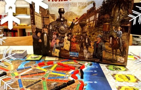 Alt text: Ticket to Ride: Legends of the West board game box displayed on a table with an illustrated game board featuring colorful train routes and player pieces. The scene is decorated with snowflake graphics, indicating a festive theme.