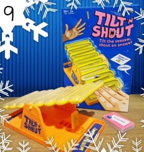 Tilt 'N Shout board game featuring a colorful seesaw design, suitable for ages 10 and up, designed for 2 or more players. The game encourages players to tilt the seesaw and shout answers, with a focus on fun and interactive gameplay. The box prominently displays instructions and includes a card for guessing types of cheese, such as cheddar.