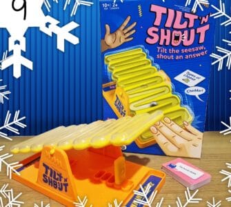 Tilt 'N Shout board game featuring a colorful seesaw design, suitable for ages 10 and up, designed for 2 or more players. The game encourages players to tilt the seesaw and shout answers, with a focus on fun and interactive gameplay. The box prominently displays instructions and includes a card for guessing types of cheese, such as cheddar.