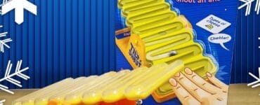 Tilt 'N Shout board game featuring a colorful seesaw design, suitable for ages 10 and up, designed for 2 or more players. The game encourages players to tilt the seesaw and shout answers, with a focus on fun and interactive gameplay. The box prominently displays instructions and includes a card for guessing types of cheese, such as cheddar.