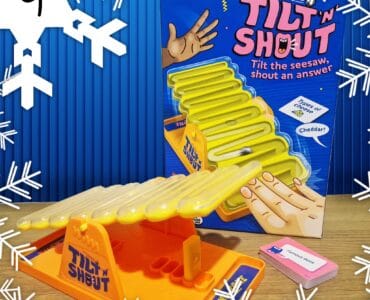 Tilt 'N Shout board game featuring a colorful seesaw design, suitable for ages 10 and up, designed for 2 or more players. The game encourages players to tilt the seesaw and shout answers, with a focus on fun and interactive gameplay. The box prominently displays instructions and includes a card for guessing types of cheese, such as cheddar.