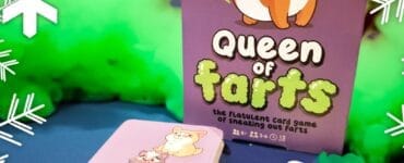Colorful image of the "Queen of Farts" card game by Professor Puzzle Games, featuring a playful corgi on the box and illustrated animal characters on the cards. The game promotes fun and laughter for ages 8 and up, designed for 2 to 6 players. Background includes green smoke effects, enhancing the playful theme.
