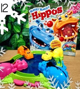 Colorful game setup of Hungry Hungry Hippos featuring pink, orange, yellow, and blue hippos around a water-filled pond, with the game box displayed in the background, all set against a lush green foliage backdrop.