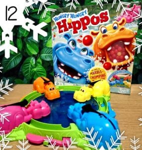 Colorful game setup of Hungry Hungry Hippos featuring pink, orange, yellow, and blue hippos around a water-filled pond, with the game box displayed in the background, all set against a lush green foliage backdrop.