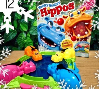 Colorful game setup of Hungry Hungry Hippos featuring pink, orange, yellow, and blue hippos around a water-filled pond, with the game box displayed in the background, all set against a lush green foliage backdrop.