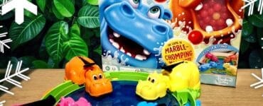 Colorful game setup of Hungry Hungry Hippos featuring pink, orange, yellow, and blue hippos around a water-filled pond, with the game box displayed in the background, all set against a lush green foliage backdrop.