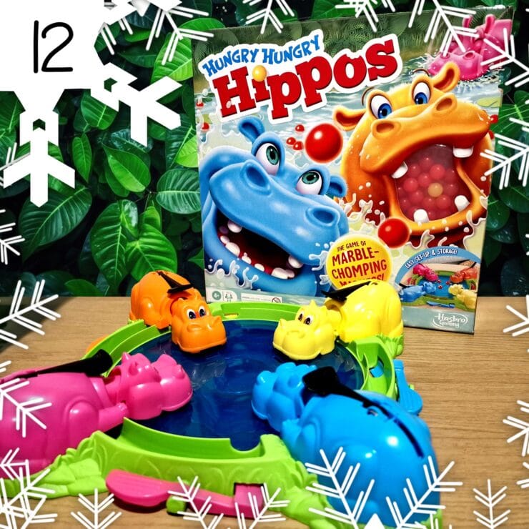 Colorful game setup of Hungry Hungry Hippos featuring pink, orange, yellow, and blue hippos around a water-filled pond, with the game box displayed in the background, all set against a lush green foliage backdrop.