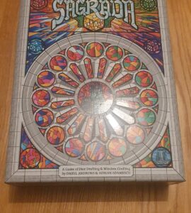 Board game Sagrada box featuring colorful stained glass window design, showcasing the title and subtitle "A Game of Dice Drafting & Window Crafting" by Daryl Andrews and Adrian Adamescu, placed on a wooden surface.