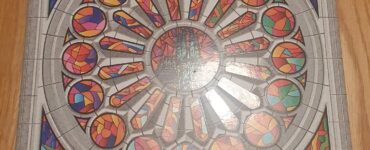 Board game Sagrada box featuring colorful stained glass window design, showcasing the title and subtitle "A Game of Dice Drafting & Window Crafting" by Daryl Andrews and Adrian Adamescu, placed on a wooden surface.