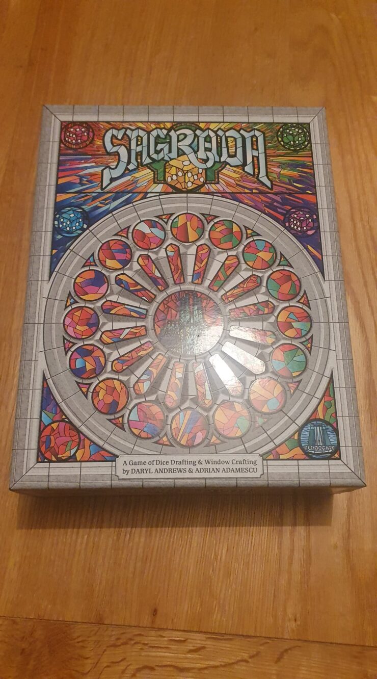 Board game Sagrada box featuring colorful stained glass window design, showcasing the title and subtitle "A Game of Dice Drafting & Window Crafting" by Daryl Andrews and Adrian Adamescu, placed on a wooden surface.