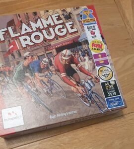 Flamme Rouge board game box featuring a vintage cycling theme, with vibrant artwork of cyclists racing. The box displays multiple awards, including the 2018 As d'Or and Mensa Select, highlighting its popularity and acclaim. The game is designed for family and strategy enthusiasts.