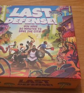 Board game box of "Last Defense!" featuring vibrant artwork of characters defending a city from alien invaders, with the tagline "You have 20 minutes to save the city!" and designed for 2-8 players.