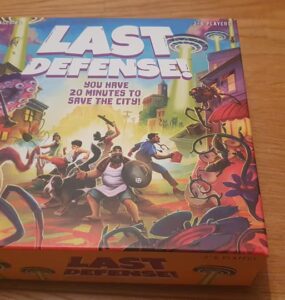 Board game box of "Last Defense!" featuring vibrant artwork of characters defending a city from alien invaders, with the tagline "You have 20 minutes to save the city!" and designed for 2-8 players.