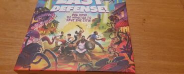 Board game box of "Last Defense!" featuring vibrant artwork of characters defending a city from alien invaders, with the tagline "You have 20 minutes to save the city!" and designed for 2-8 players.