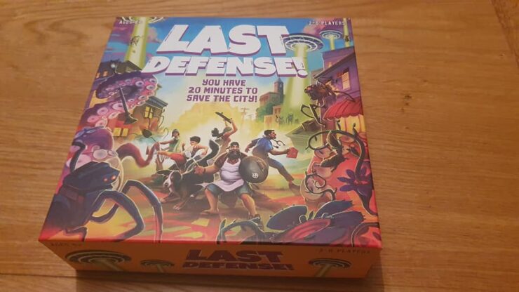 Board game box of "Last Defense!" featuring vibrant artwork of characters defending a city from alien invaders, with the tagline "You have 20 minutes to save the city!" and designed for 2-8 players.