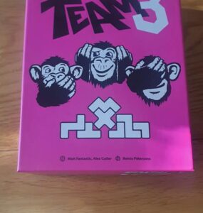 Colorful game box for "Team 3," featuring three cartoon monkeys with playful expressions, set against a bright pink background. The design includes the game's title and logos of the creators, Matt Fantastic, Alex Cutler, and Reinis Pētersons. Perfect for board game enthusiasts.