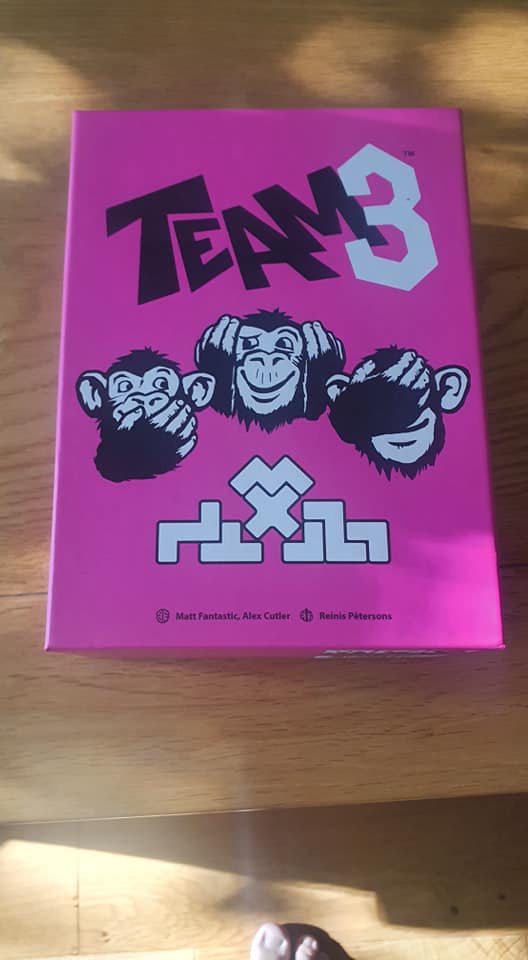 Colorful game box for "Team 3," featuring three cartoon monkeys with playful expressions, set against a bright pink background. The design includes the game's title and logos of the creators, Matt Fantastic, Alex Cutler, and Reinis Pētersons. Perfect for board game enthusiasts.